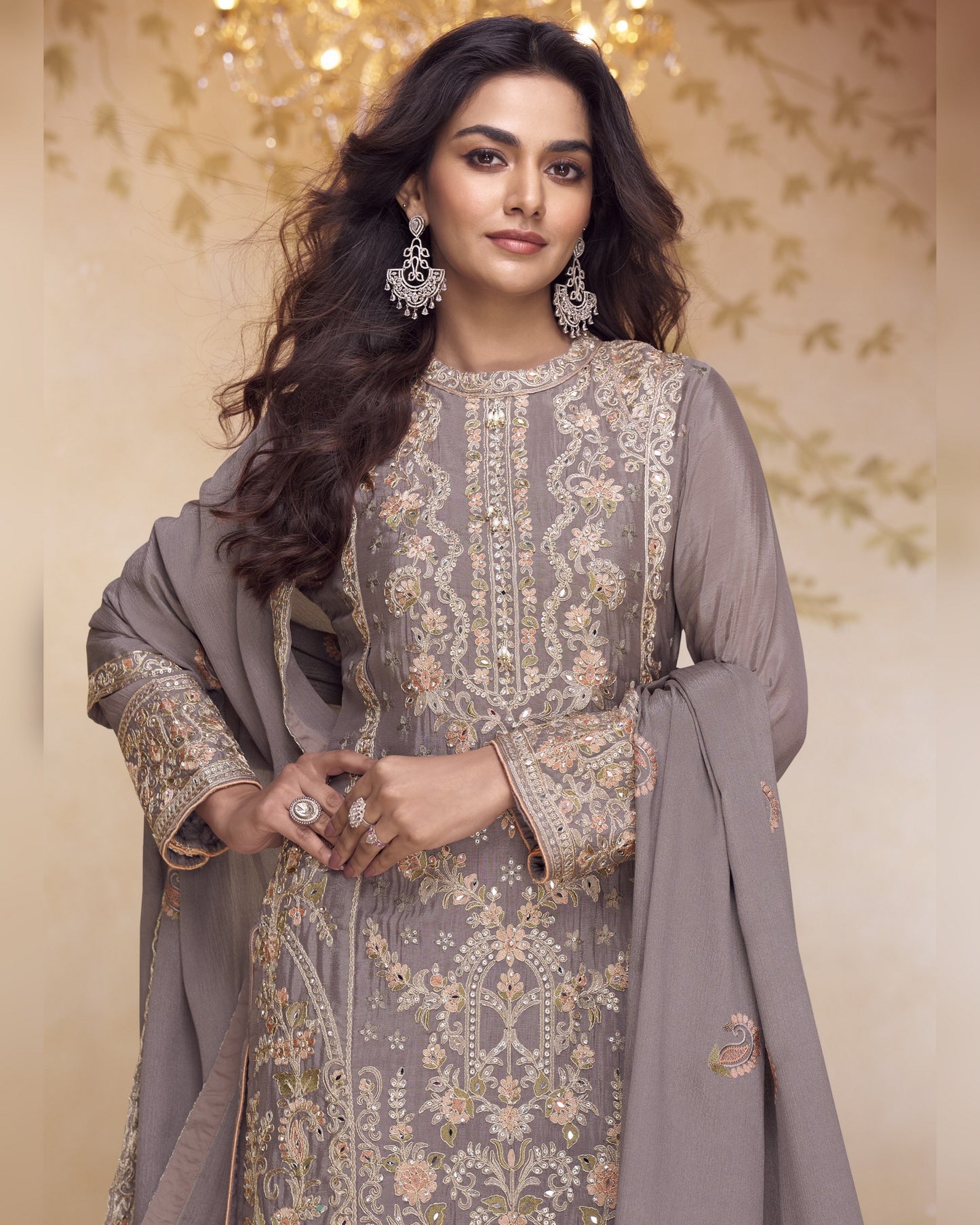 Woman wearing a gray embroidered traditional outfit with a matching dupatta against a decorative background.