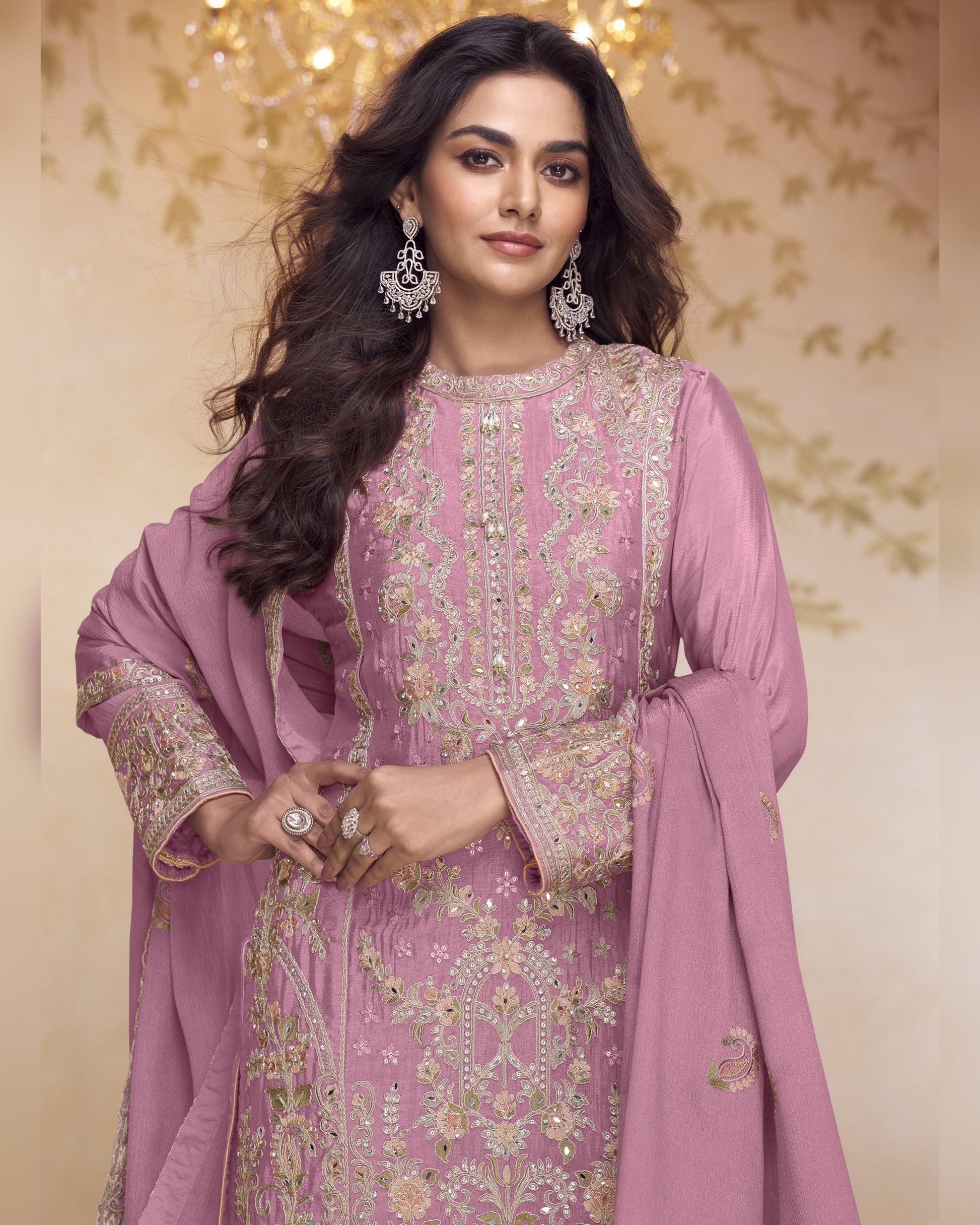 Woman wearing a pink embroidered traditional outfit with a matching dupatta against a decorative background.