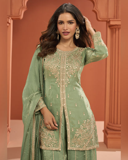 Woman wearing a green embroidered traditional outfit with a matching dupatta against an orange architectural background.