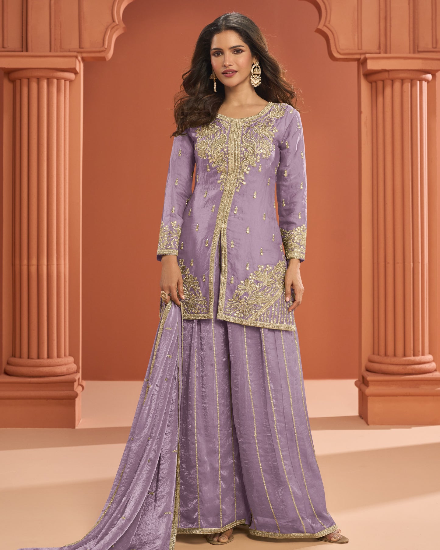 Woman wearing a traditional lavender outfit with gold embroidery against an architectural background.