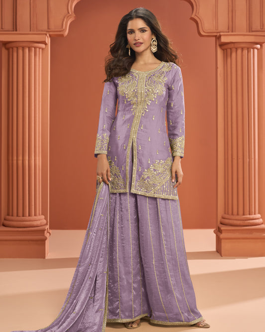 Woman wearing a traditional lavender outfit with gold embroidery against an architectural background.