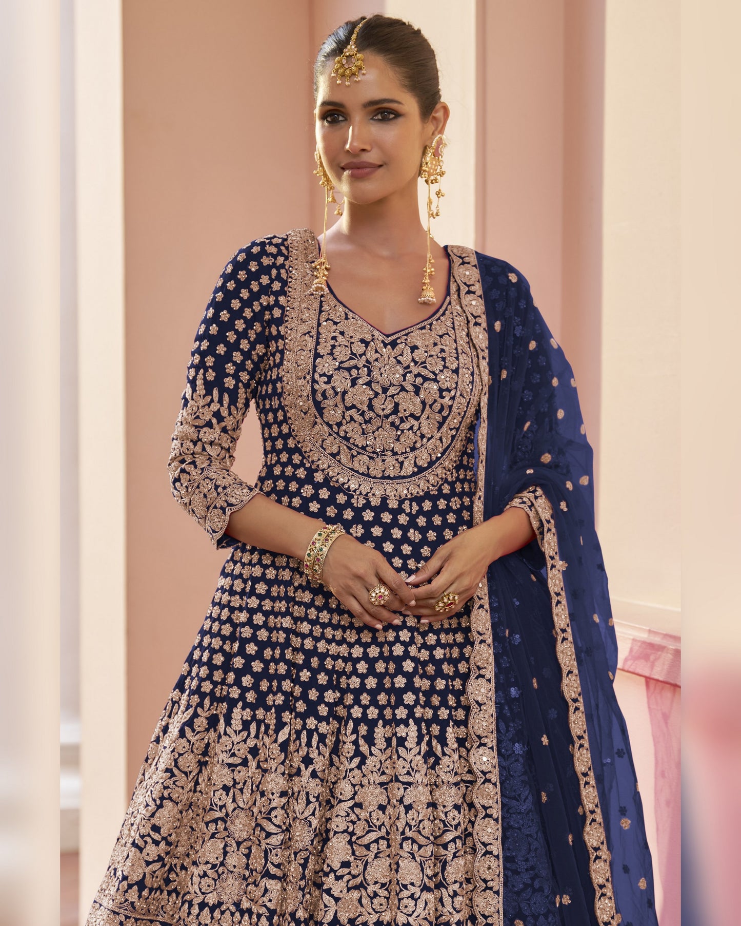 Woman wearing a traditional embroidered outfit with a blue dupatta against a light pink background
