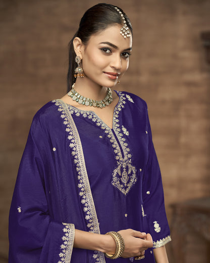 Woman wearing a traditional purple outfit with intricate embroidery and jewelry against a brown background