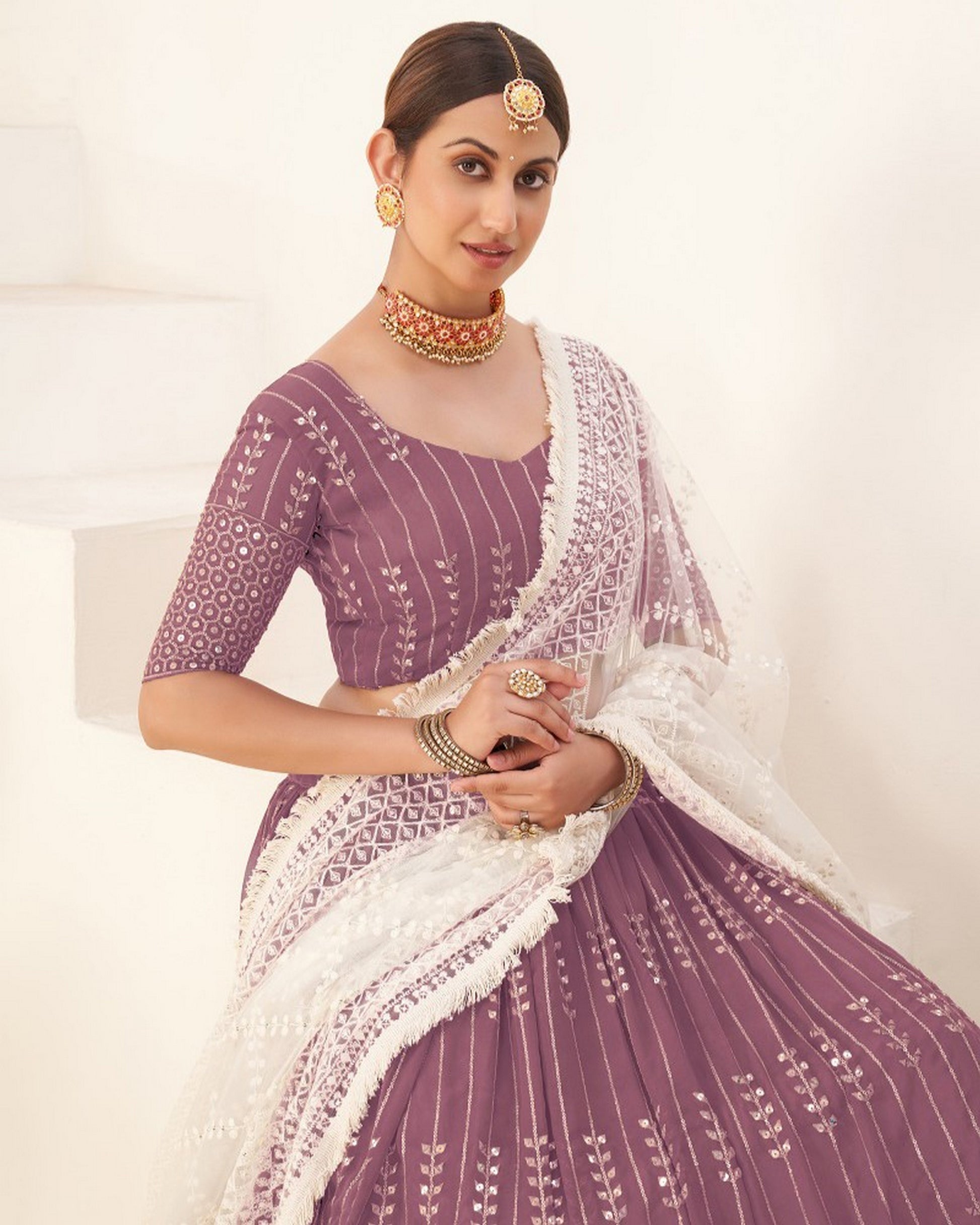 Woman wearing a traditional purple and white embroidered outfit with jewelry on a light background