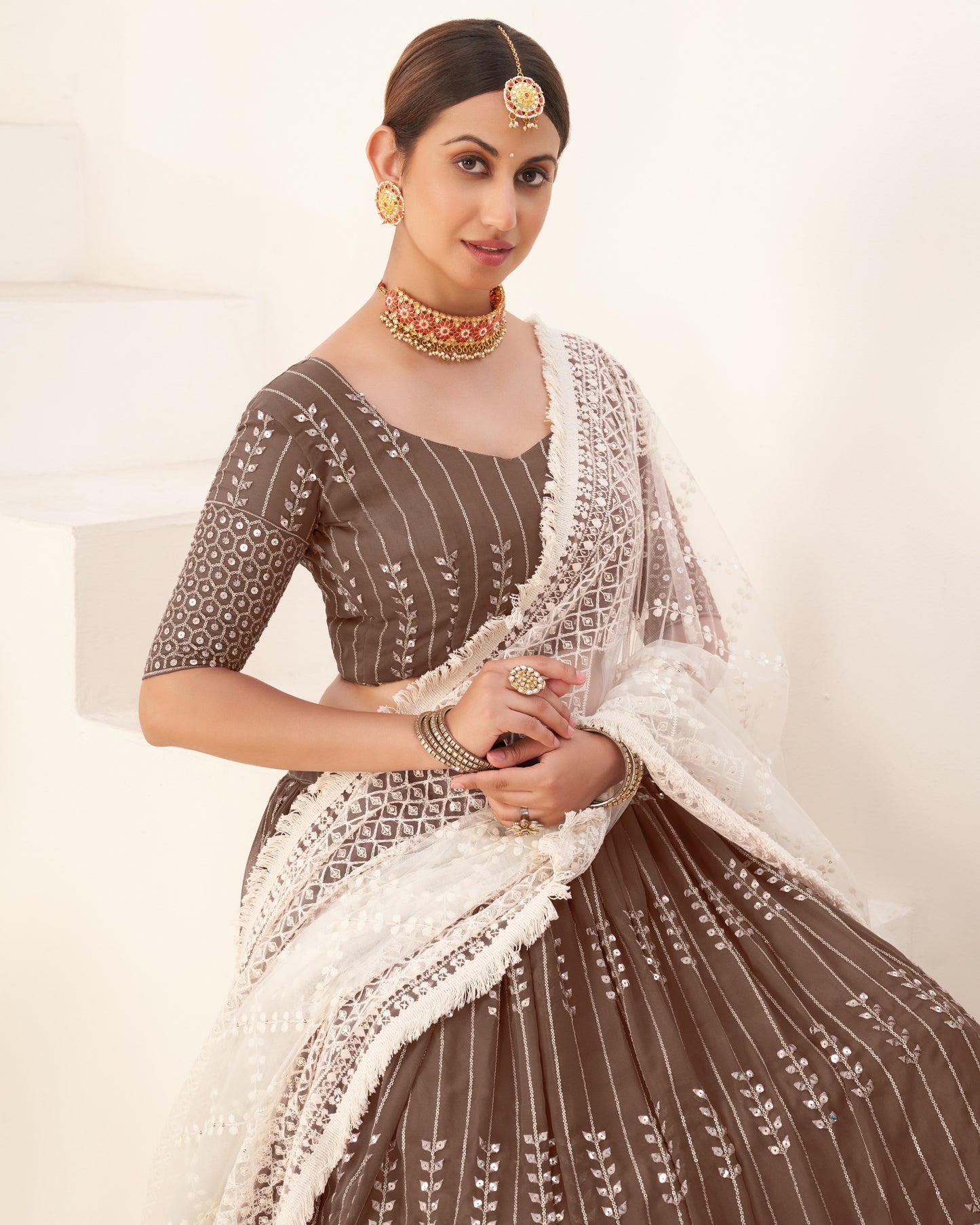 Woman wearing a traditional brown and white embroidered outfit with jewelry on a light background