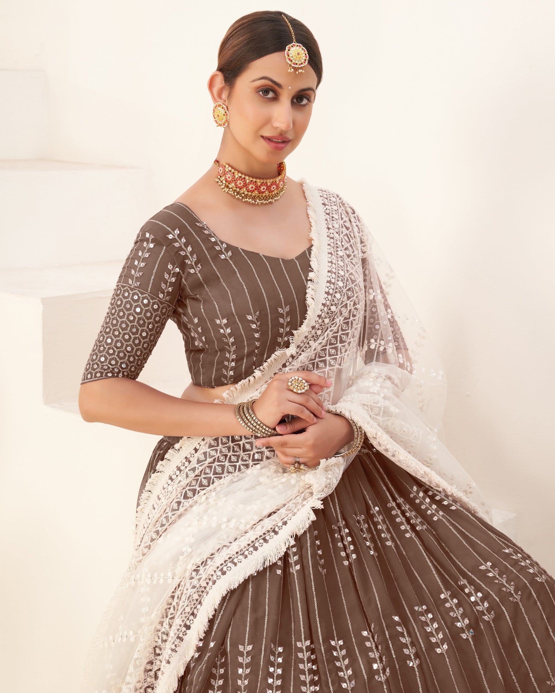 Woman wearing a traditional brown and white embroidered outfit with jewelry on a light background