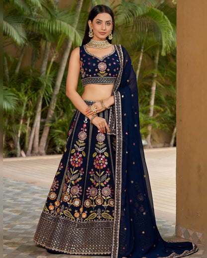 Woman in a traditional navy blue embroidered outfit standing outdoors with greenery in the background