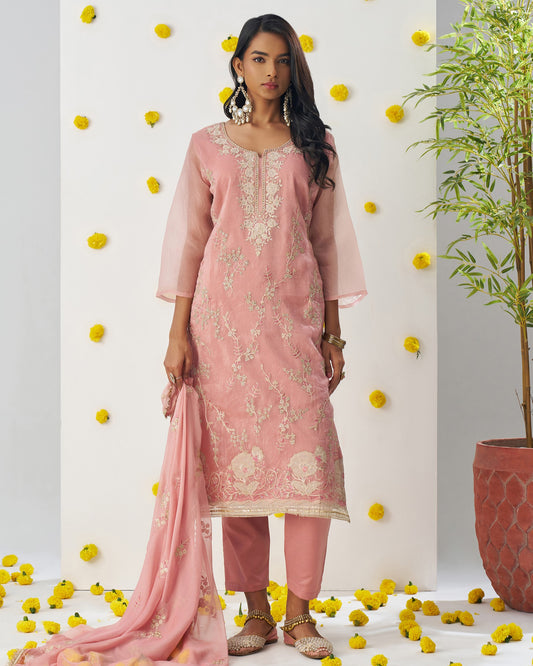 Woman in a pink traditional outfit with floral patterns standing against a decorated wall with yellow flowers and a plant.