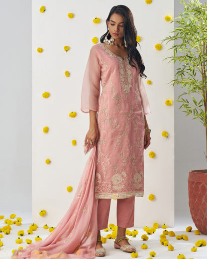 Woman in a pink embroidered outfit standing among yellow flowers and a plant.