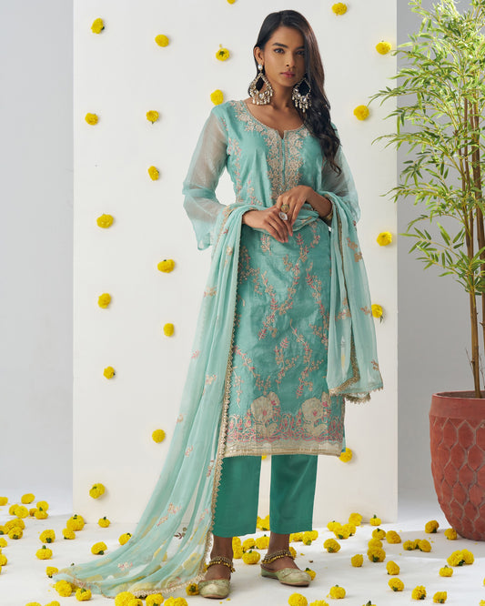 Woman in a teal traditional outfit with a matching dupatta, standing on yellow flowers with a plant in the background.