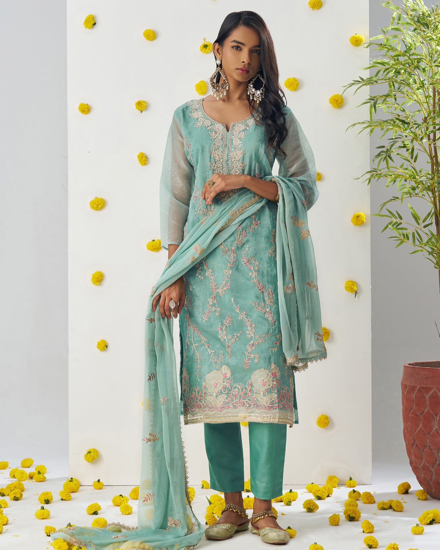 Woman in a light blue embroidered outfit with yellow flowers and a plant in the background