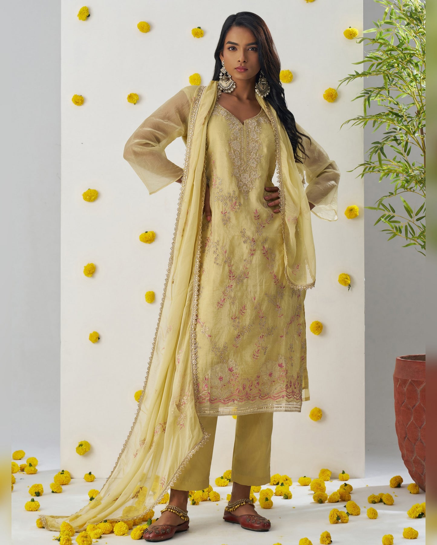 Woman in a light yellow traditional outfit with a dupatta, standing against a decorated wall with yellow flowers.