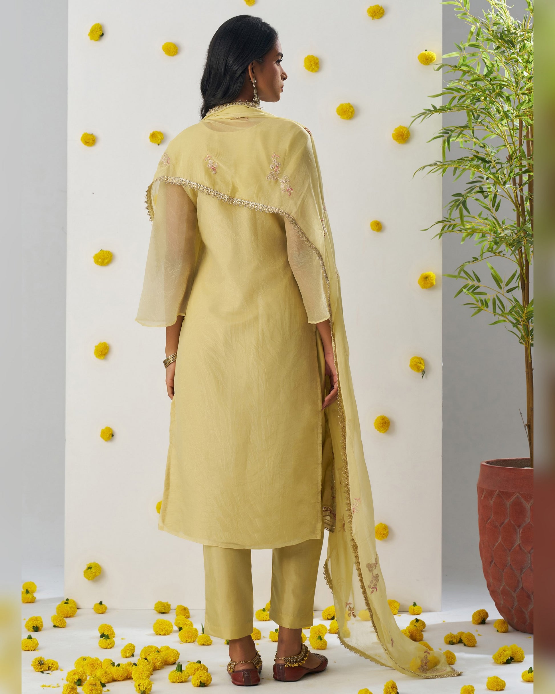 Woman in a yellow traditional outfit with a dupatta, standing against a decorated wall with yellow flowers and a plant.