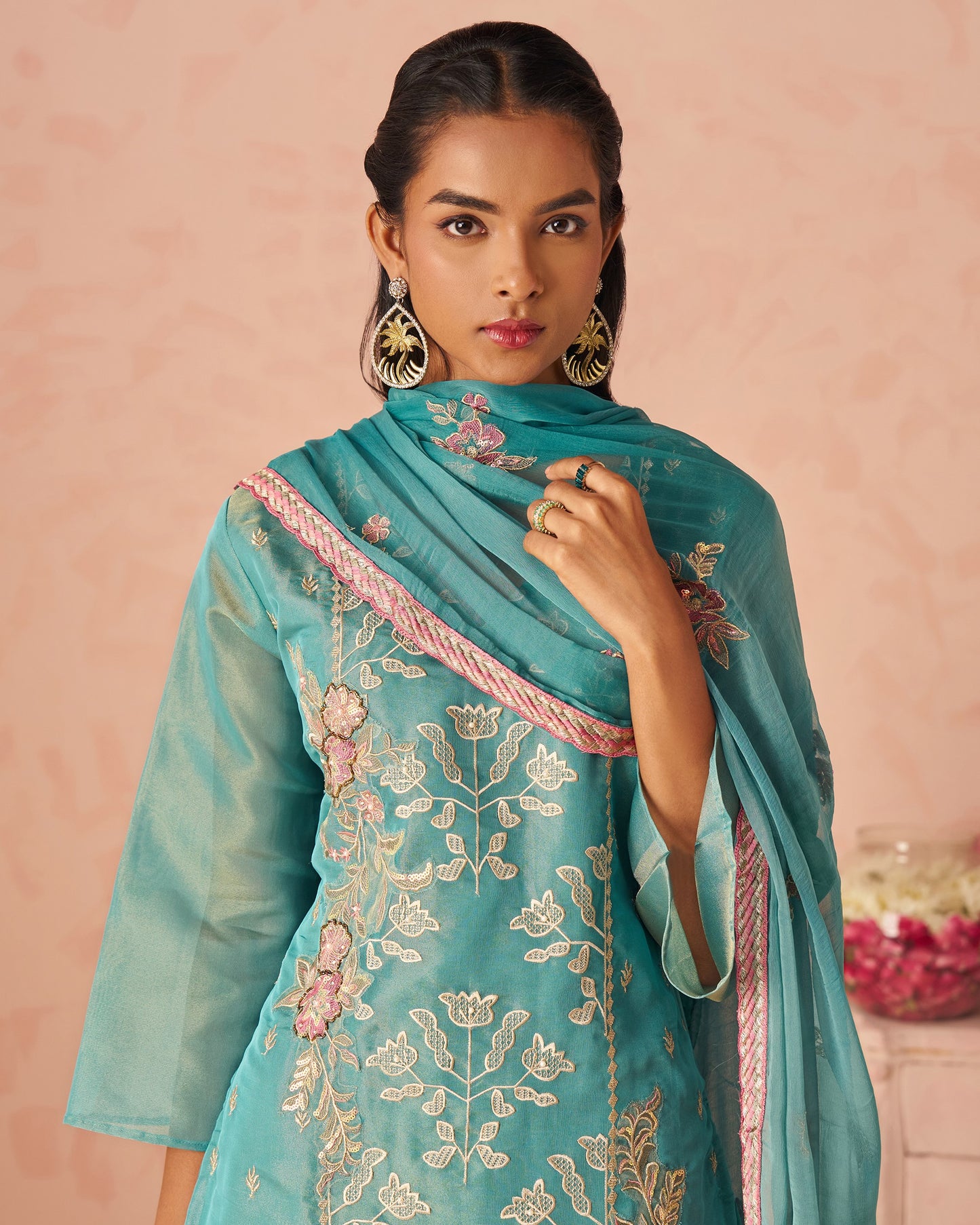 Woman wearing a turquoise embroidered outfit with a pink border against a pink background