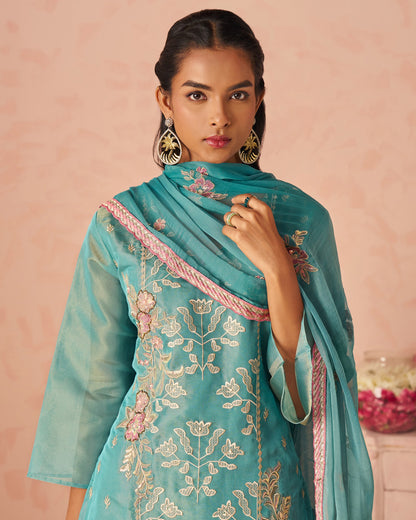 Woman wearing a turquoise embroidered outfit with a pink border against a pink background