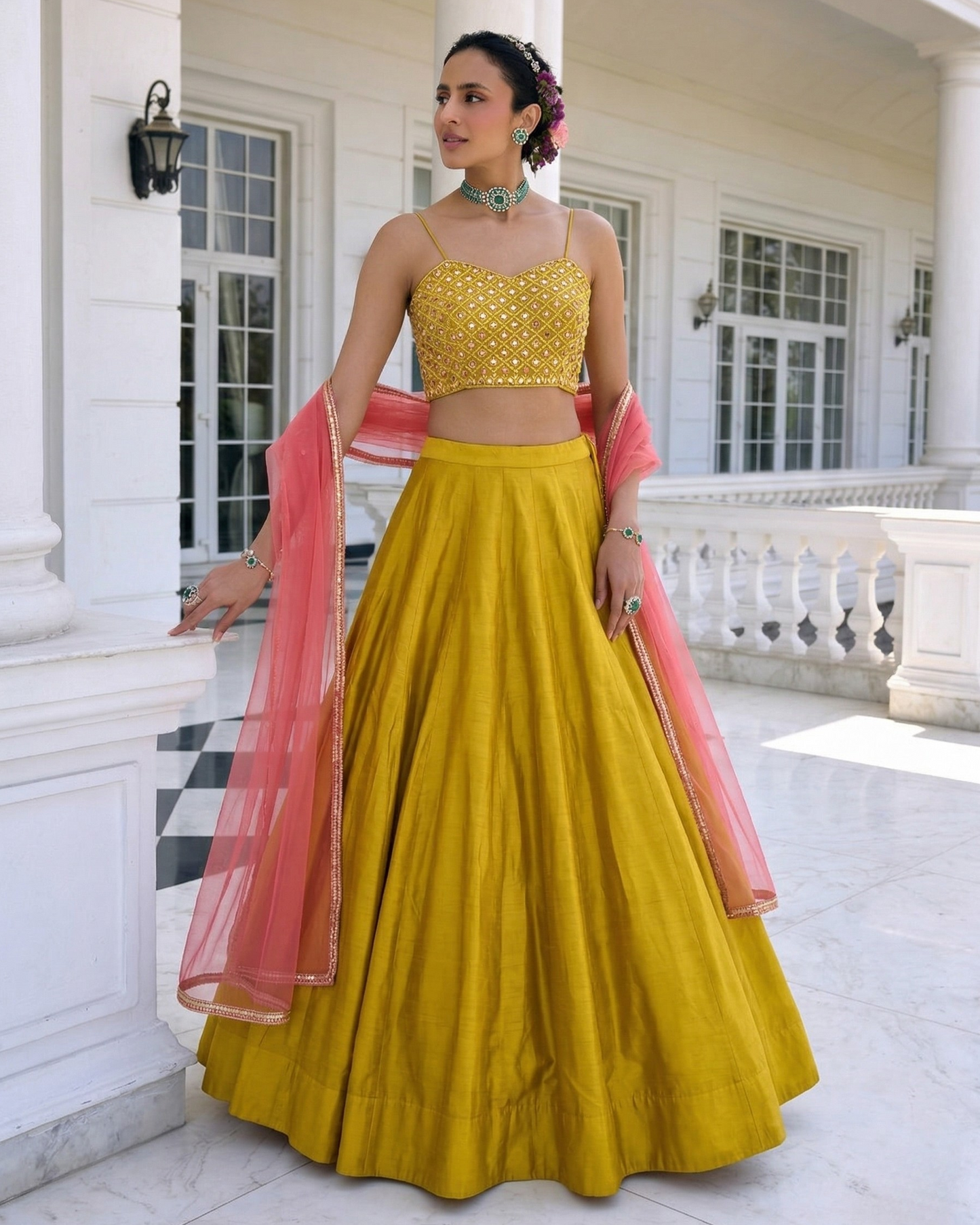 Woman in a yellow traditional outfit with pink dupatta standing in front of a white building.