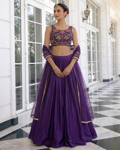 Woman wearing a purple traditional outfit with a colorful top in front of a white building.