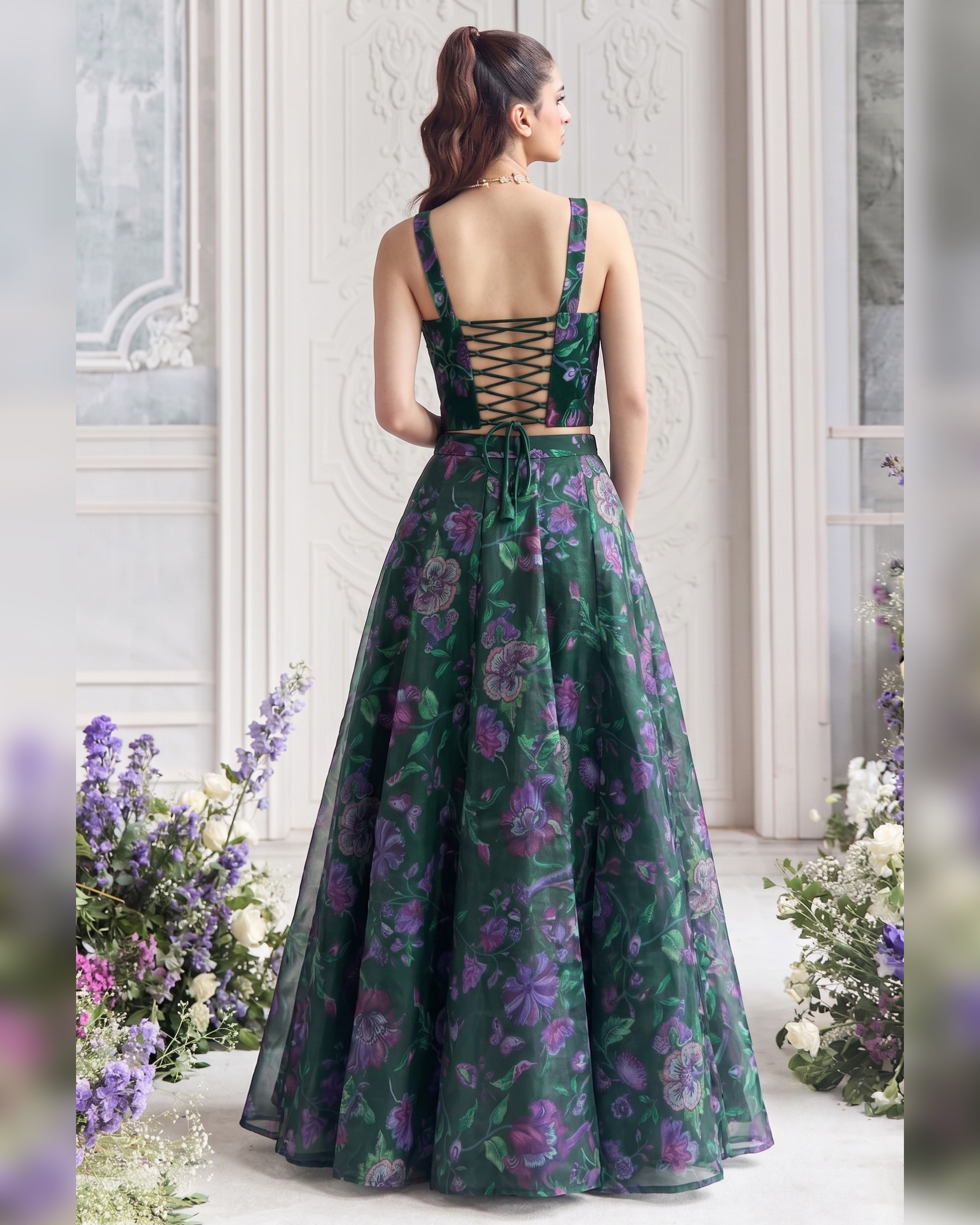 A woman stands with her back to the camera wearing a formal floor-length gown with a dark green floral print, a lace-up open back, and a full skirt, posed in an elegant indoor setting with floral arrangements.