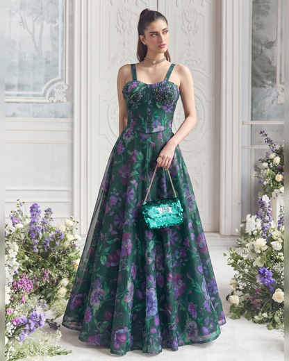 A woman models a floor-length green evening gown with a purple and blue floral pattern, holding a shiny teal handbag in an elegant indoor setting with floral arrangements.