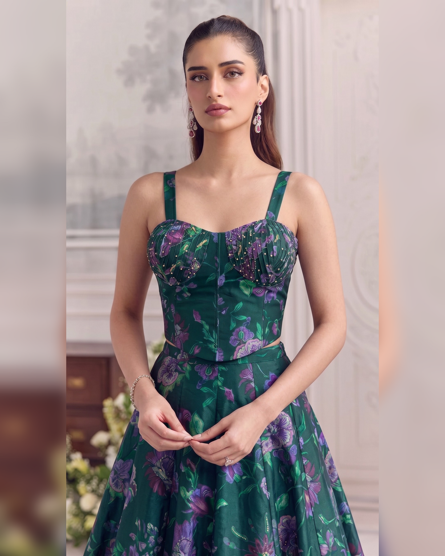 A woman modeling a dark green floral print dress with purple and pink floral motifs, featuring a structured sweetheart bodice, thin shoulder straps, and a flared skirt, set against a softly blurred indoor background.