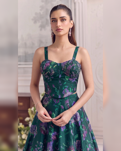 A woman modeling a dark green floral print dress with purple and pink floral motifs, featuring a structured sweetheart bodice, thin shoulder straps, and a flared skirt, set against a softly blurred indoor background.