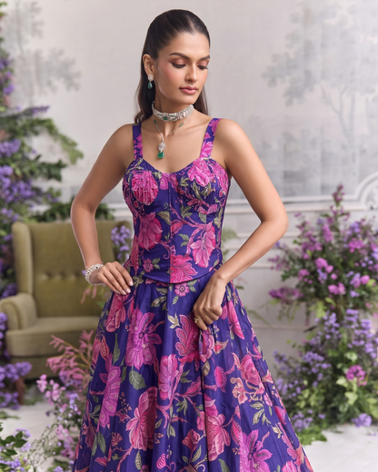 A woman models a sleeveless A-line formal gown with a purple base and a vibrant pink floral pattern, set against a softly blurred indoor garden backdrop.