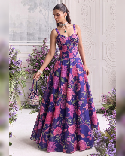 A woman models a floor-length A-line gown with a purple base and a vibrant pink floral print, standing in a decorated indoor setting with purple flowers.