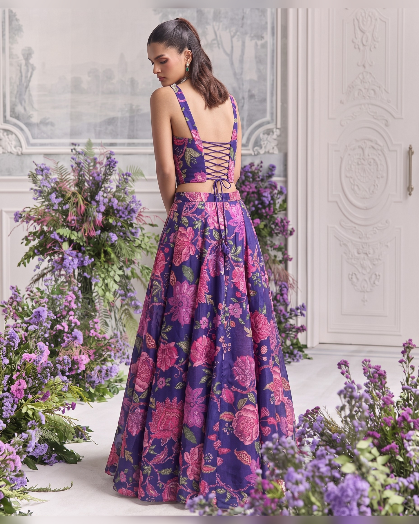 A woman stands in a garden-like interior wearing a formal A-line gown with a floral print. The dress has a purple base with pink and magenta flowers, green leaves, and a lace-up open back.