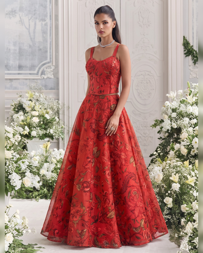 A woman stands in a grand interior wearing a voluminous red evening gown with floral embroidery and thin straps. The room has ornate white walls, large flower arrangements, and a light floor.