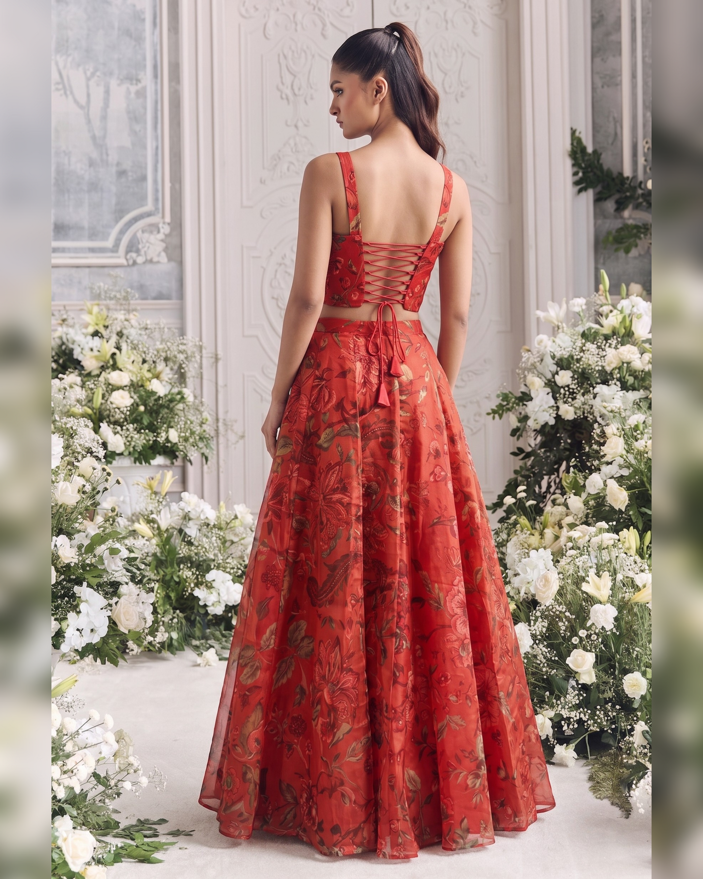A woman stands with her back to the camera wearing a red floor-length gown with a floral print and lace-up open back, posed between large white flower arrangements in an elegant indoor setting.