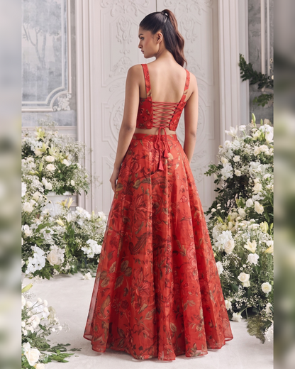 A woman stands with her back to the camera wearing a red floor-length gown with a floral print and lace-up open back, posed between large white flower arrangements in an elegant indoor setting.