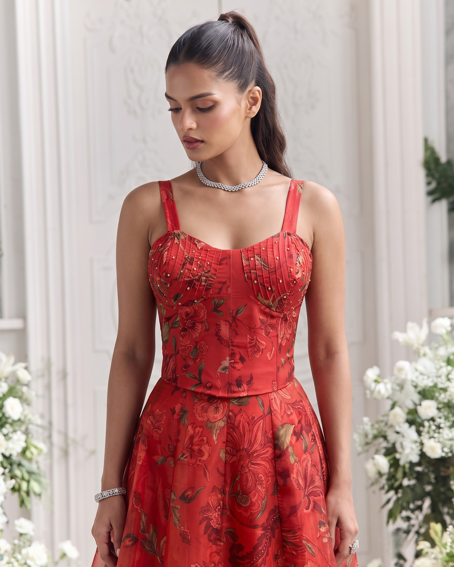 A woman wearing a red floral spaghetti-strap dress with a structured corset-style bodice and flared skirt, standing in a softly lit indoor setting with white flowers and paneled walls in the background.