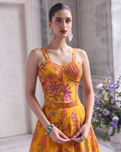 A woman models a bright yellow sleeveless dress with a pink and orange floral print, styled with ornate silver earrings and a green bracelet, set against a soft-focus classical interior background.