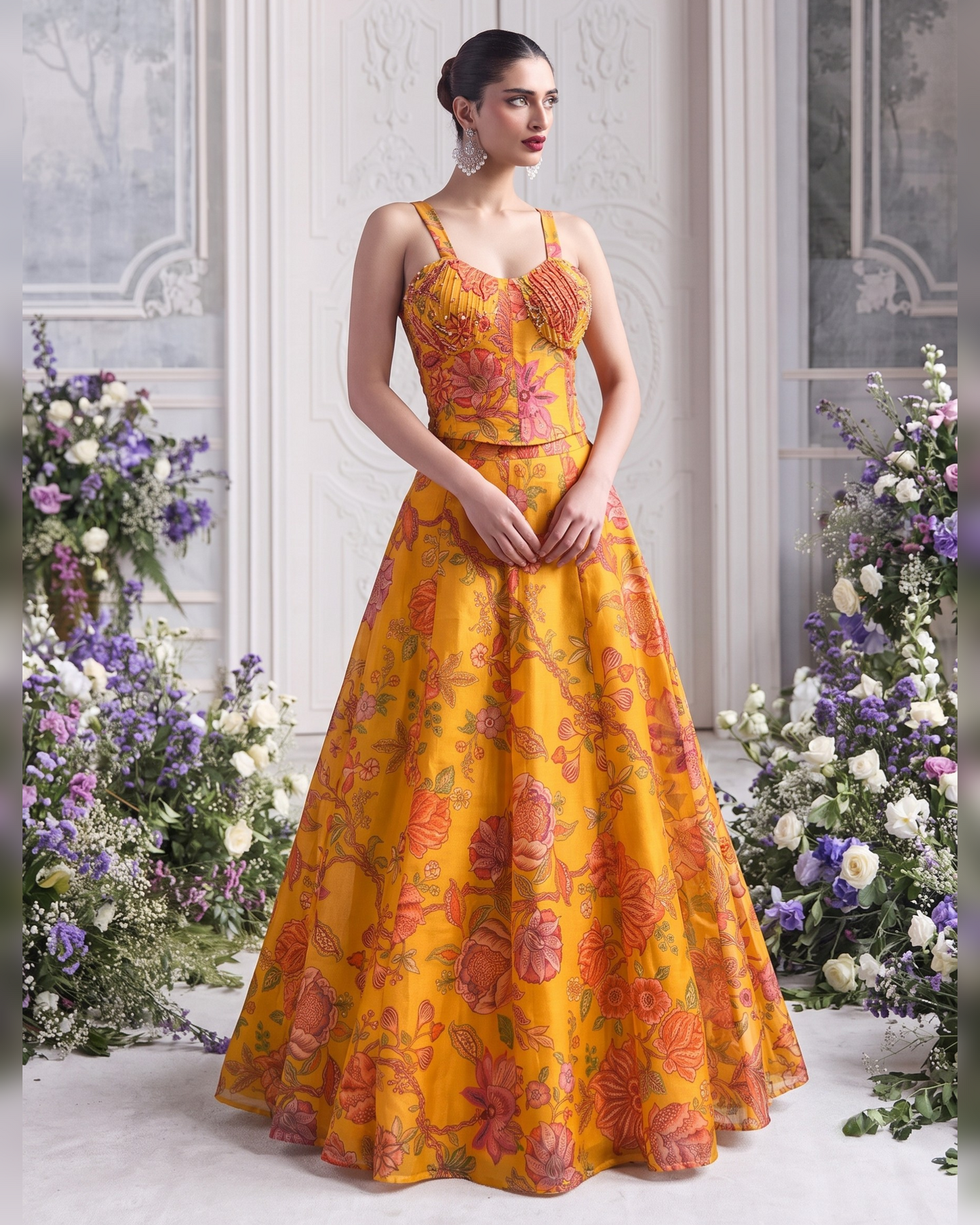 A woman models a formal ball gown with a vibrant yellow base and a pink and orange floral pattern. The dress features a structured sweetheart bodice with lace applique and a full, flowing skirt.