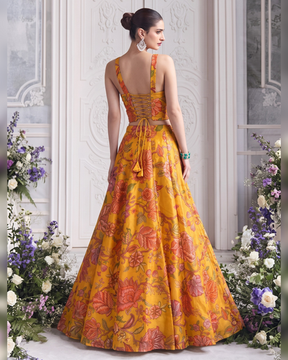 A woman stands with her back to the camera wearing a yellow and orange floral printed gown with lace-up back straps, posed in front of an ornate white paneled wall with floral arrangements on both sides.