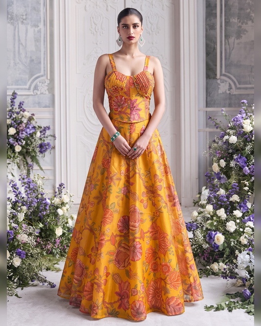 A woman stands in a grand interior wearing a bright yellow-orange floral gown with a fitted bodice and full skirt. The dress has pink and green floral patterning, and the background features white walls, decorative molding, and large arrangements of purple, white, and green flowers on both sides.