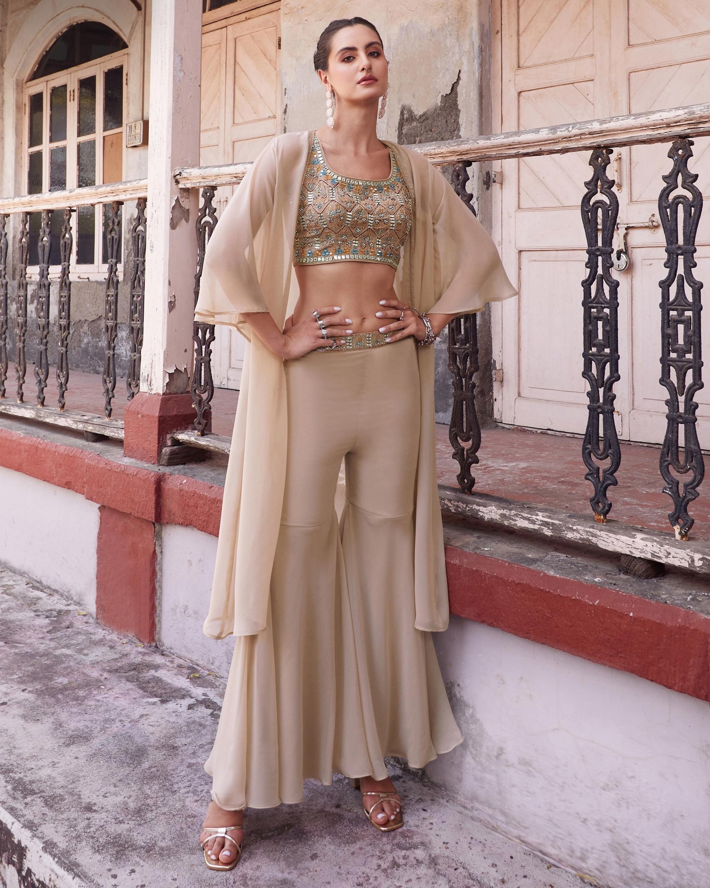 Woman in a beige outfit standing on a balcony with a textured wall and metal railing in the background.