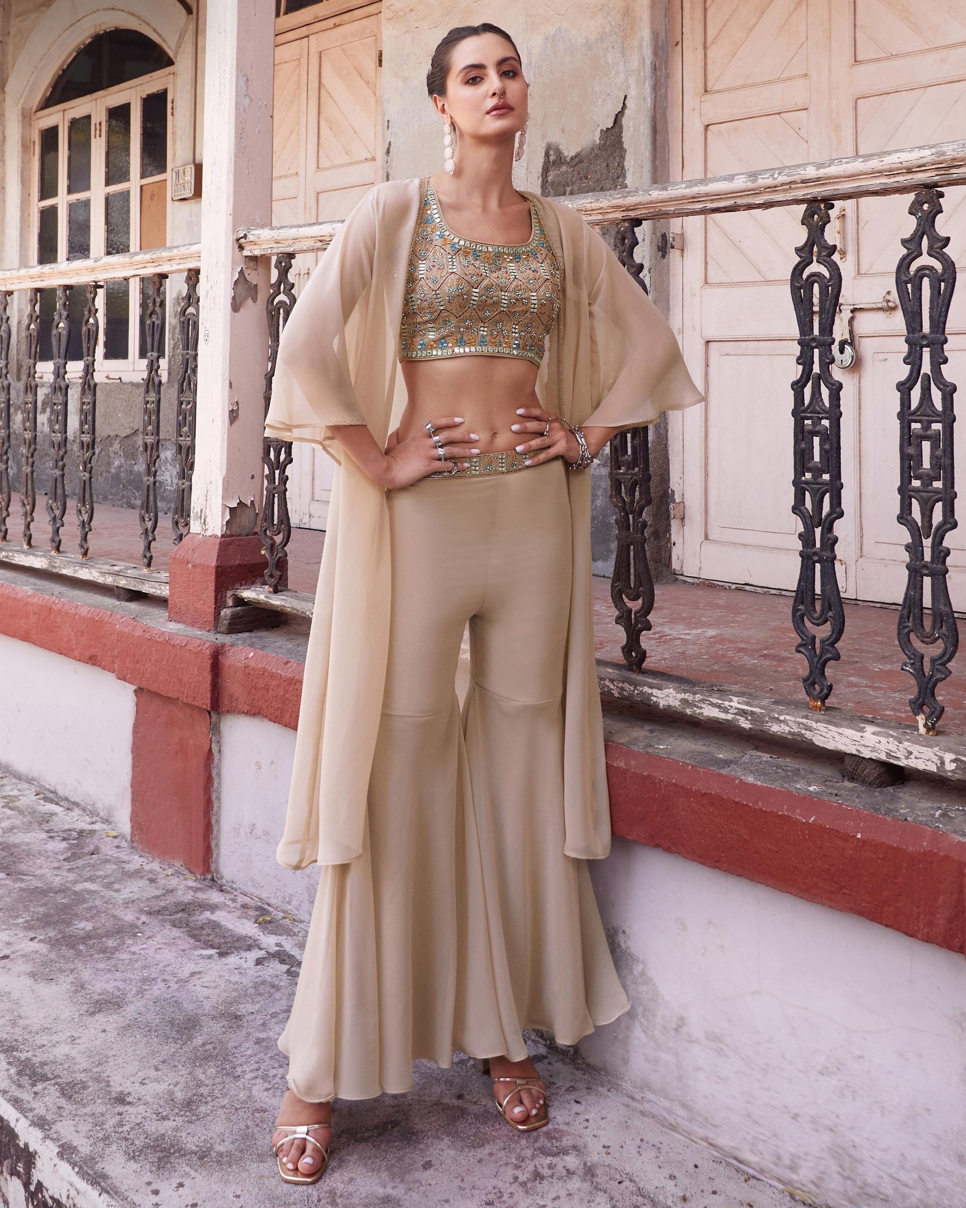 Woman in a beige outfit standing on a balcony with a textured wall and metal railing in the background.