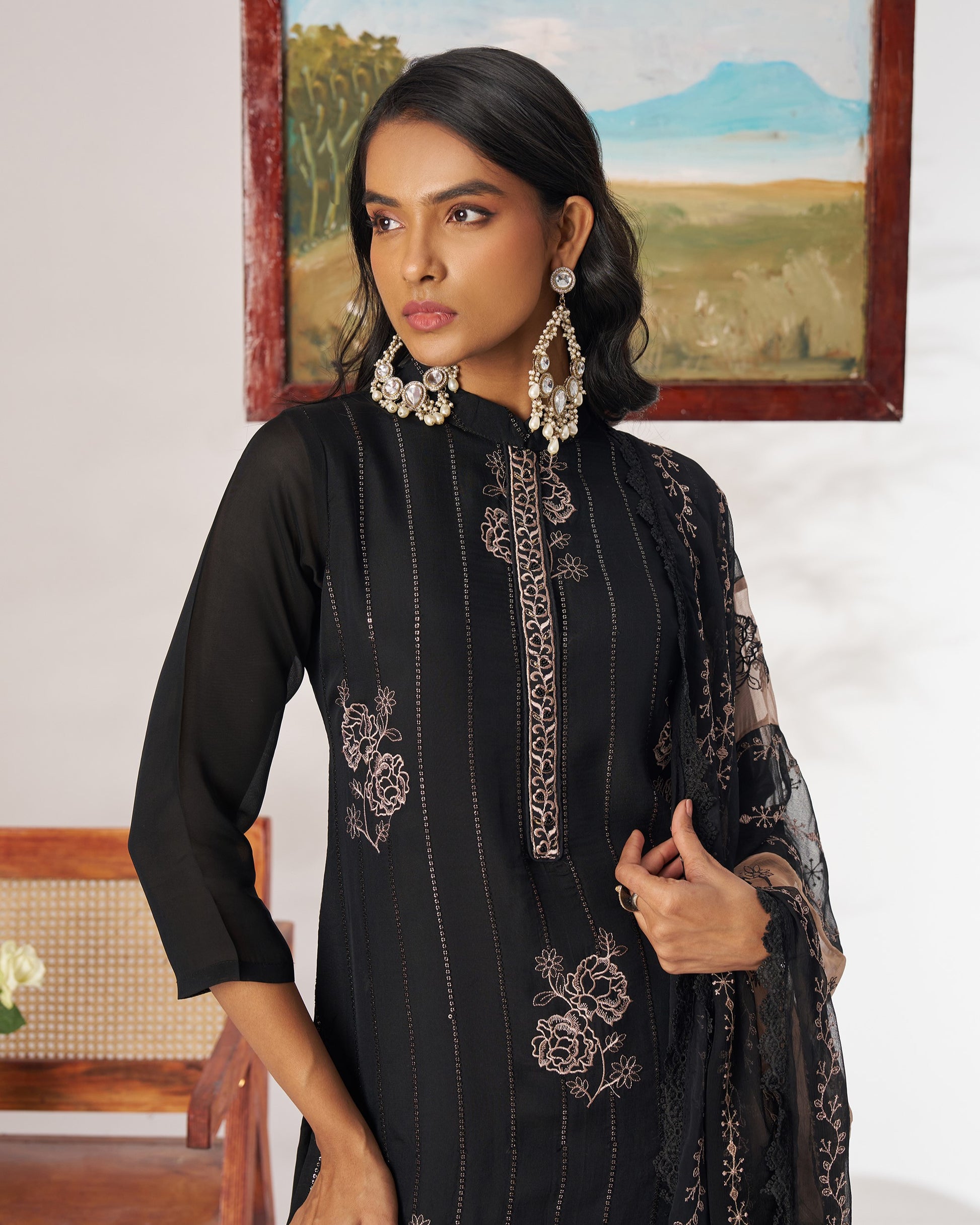 Woman wearing a black embroidered outfit with jewelry in a room with a painting on the wall.
