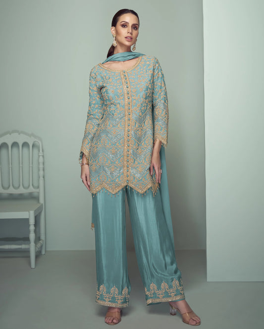 Woman wearing a light blue traditional outfit with intricate patterns in a minimalistic setting.