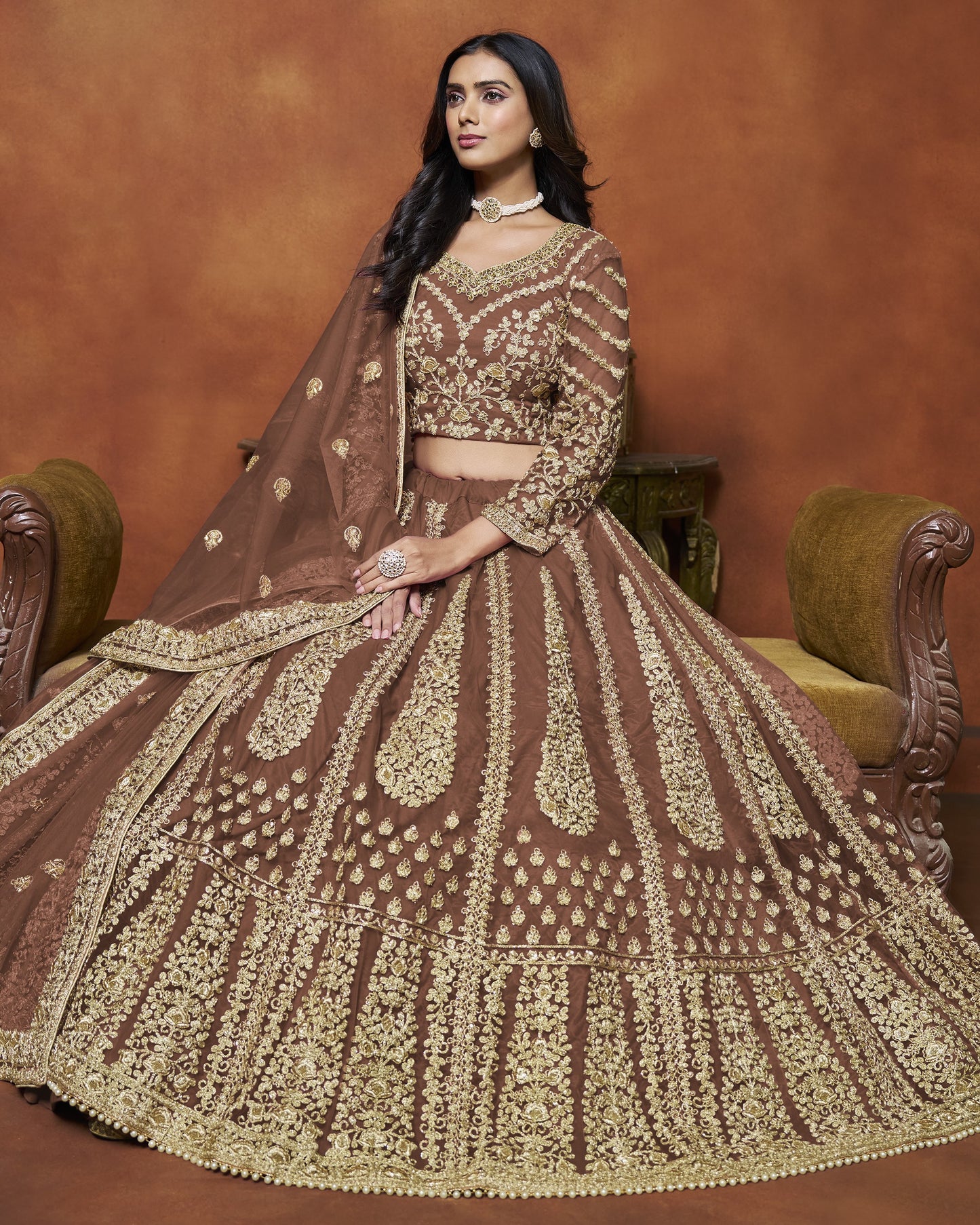 Woman in a traditional brown and gold embroidered outfit sitting on a chair against a brown background