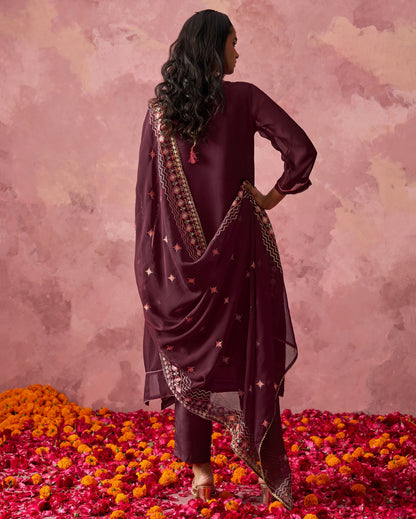 Woman in a dark purple traditional outfit with a patterned dupatta standing on flower petals against a pink floral background.