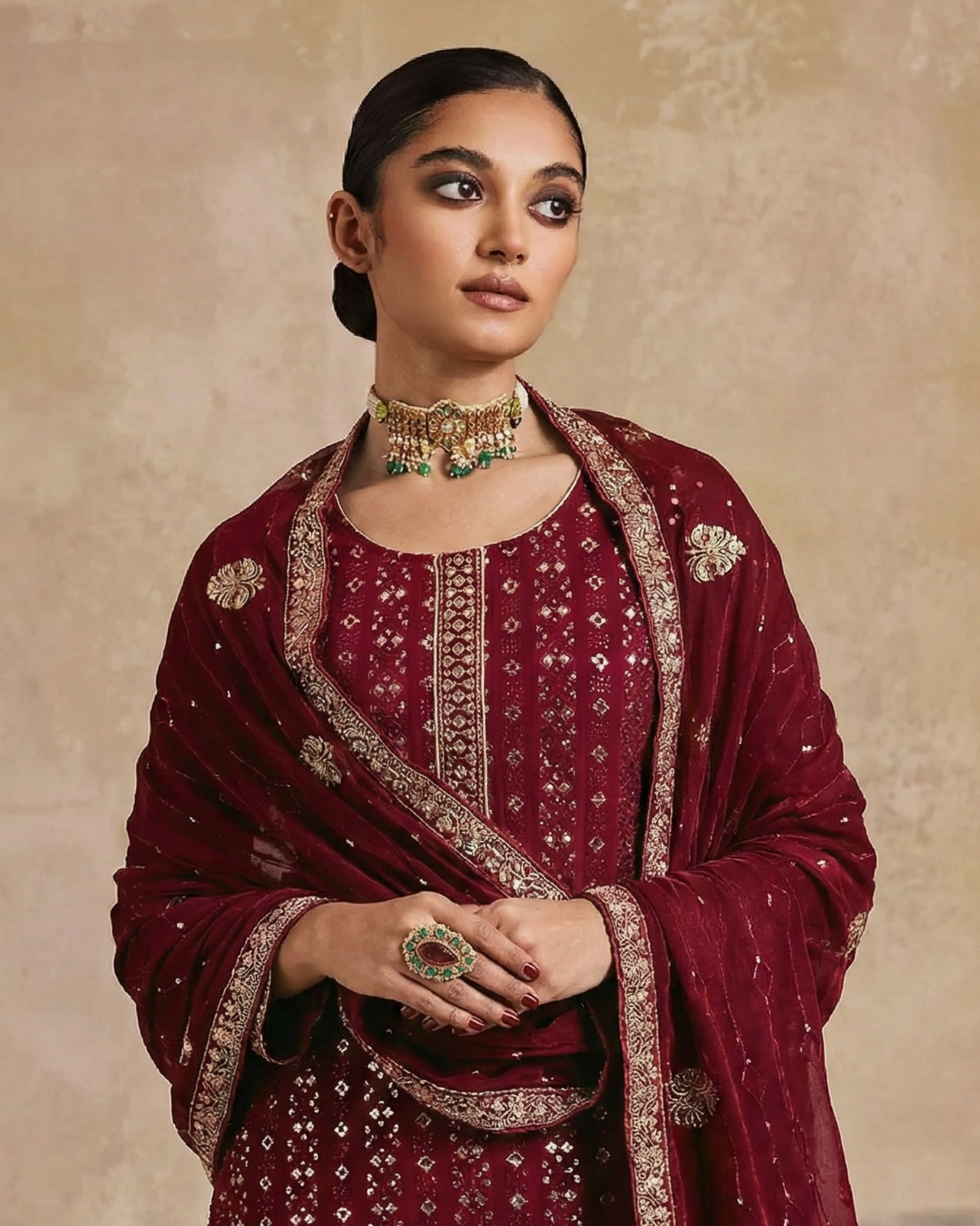 Woman wearing a maroon embroidered traditional outfit with jewelry against a beige background