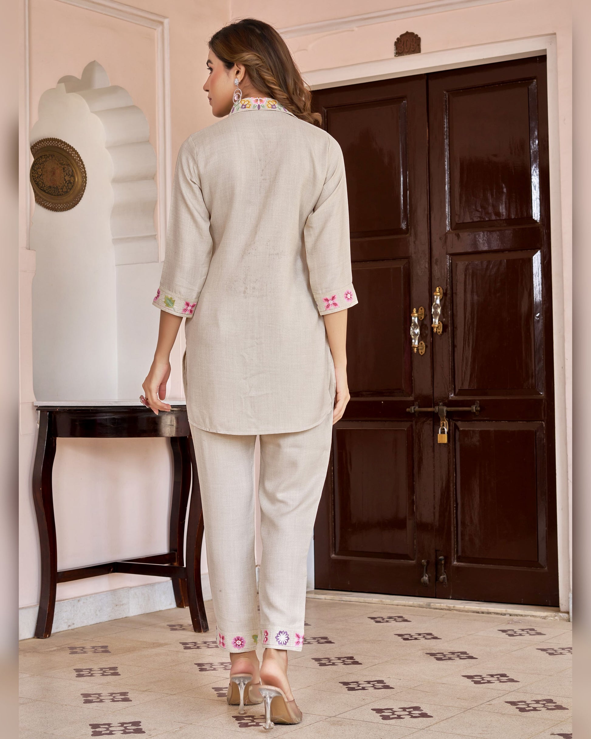 Woman in a light gray traditional outfit with pink embroidery standing in a room with a dark wooden door.
