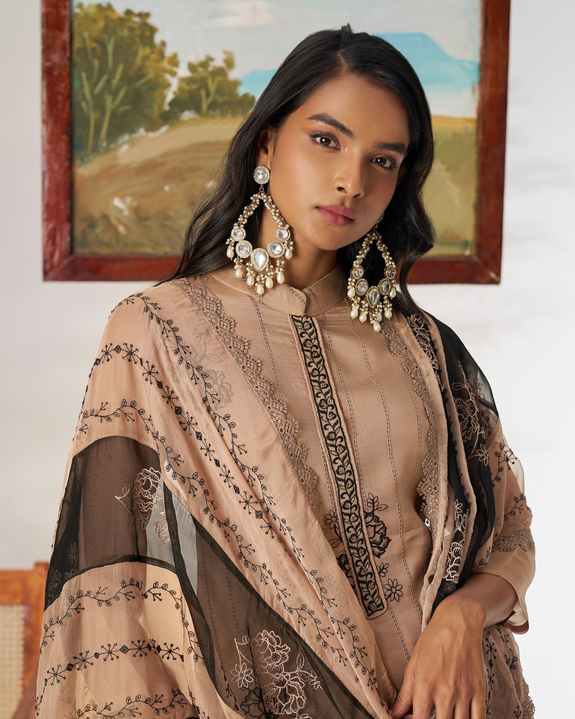 Woman wearing a beige embroidered outfit with gold earrings in a room with a painting on the wall.