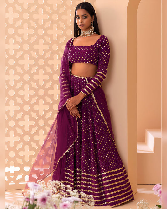 Woman in a purple traditional outfit with gold details against a decorative wall.