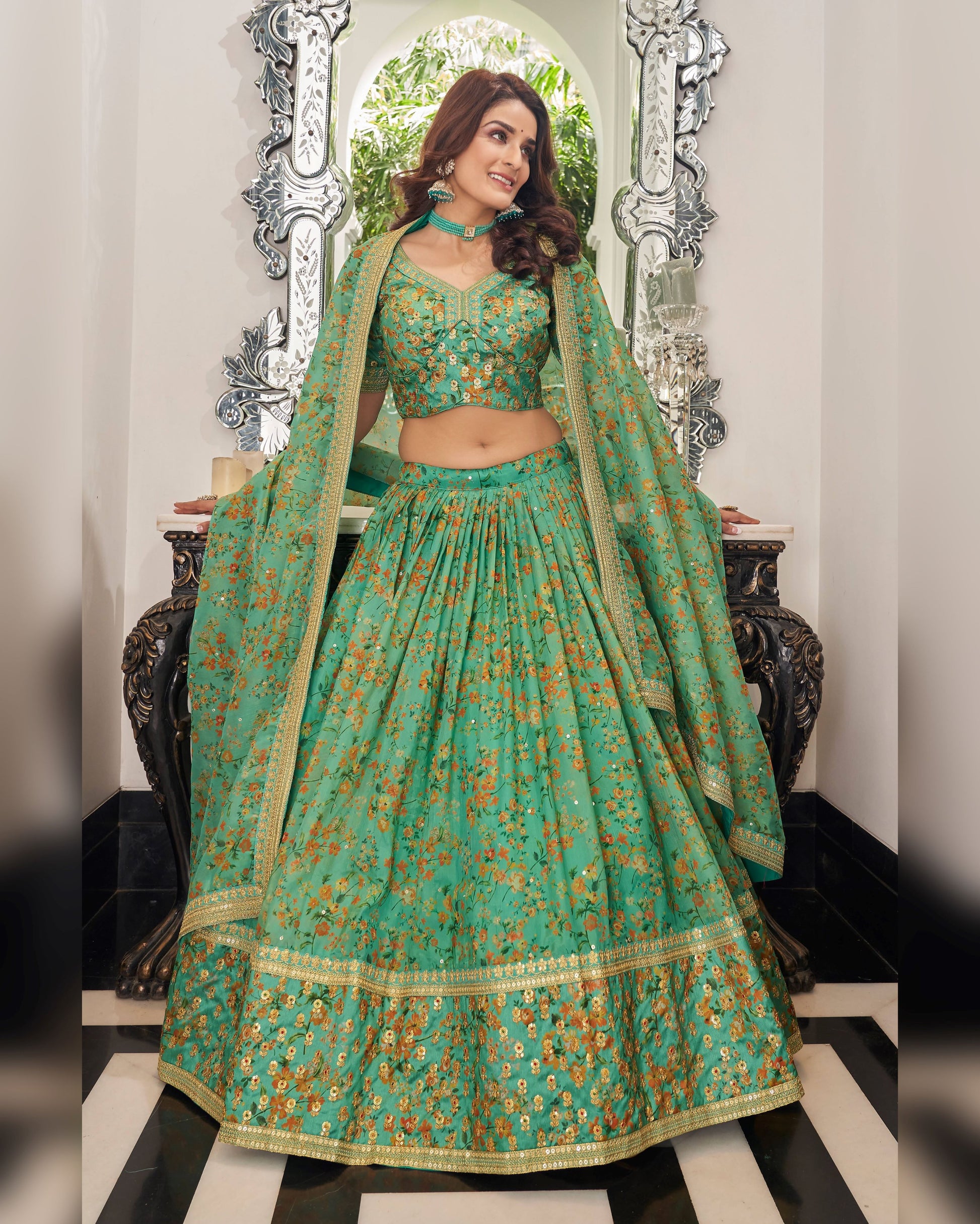 Woman in a traditional green and gold outfit with floral patterns, standing in front of a decorative mirror.