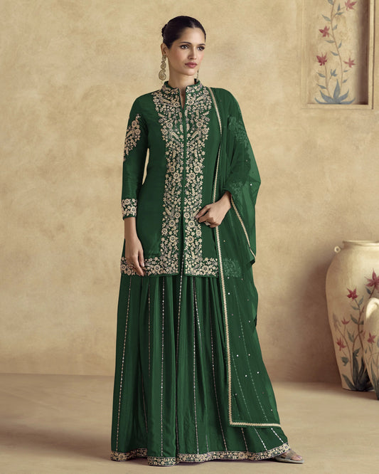 Woman wearing a green traditional outfit with embroidery in a room with a beige wall and decorative vase.