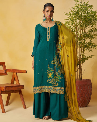 Woman in teal traditional outfit with yellow dupatta against a beige background