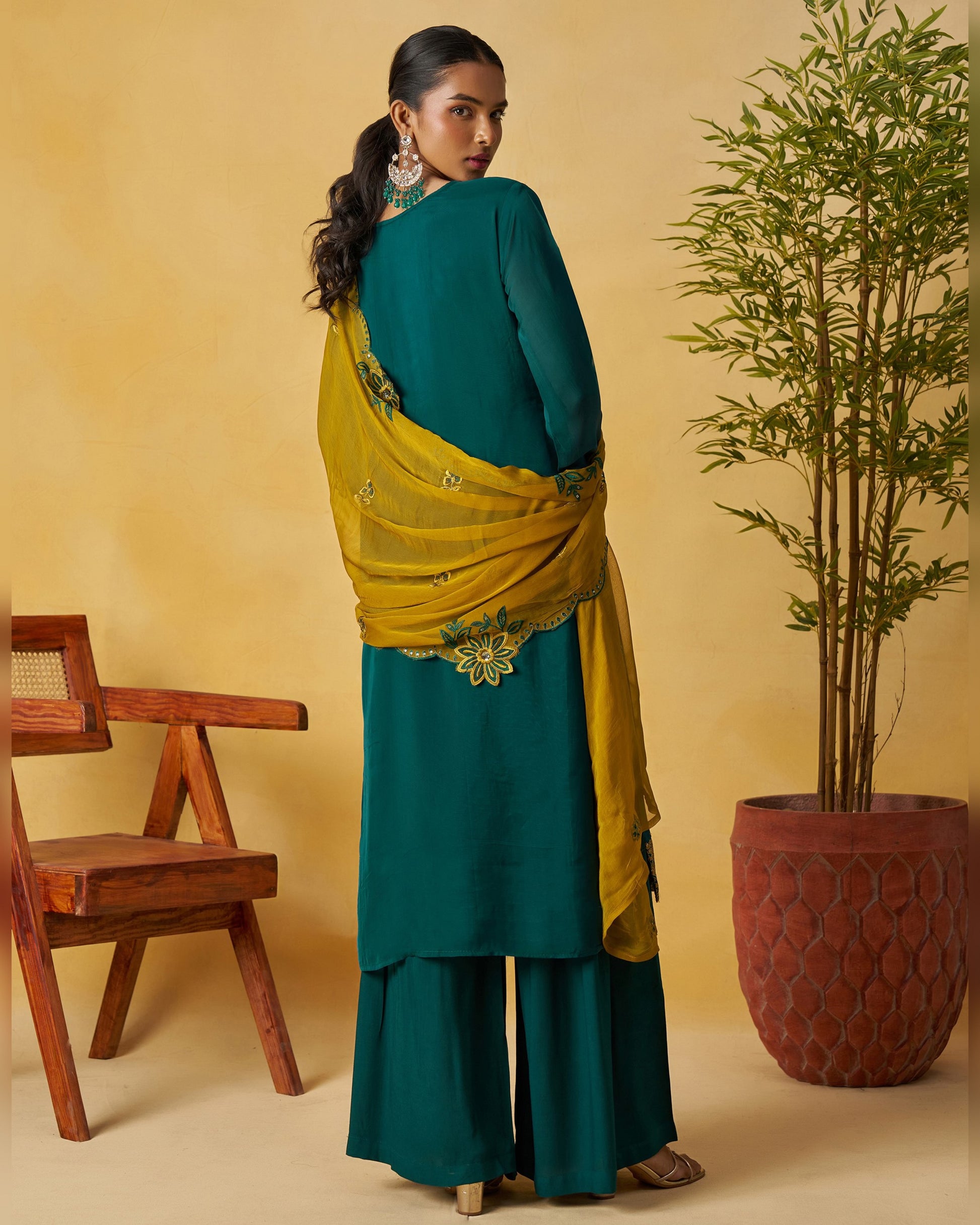 Woman in teal and yellow traditional outfit standing next to a wooden chair and potted plant on a beige background